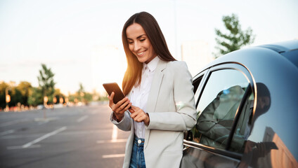 Successful smiling attractive woman in formal smart wear is using her smart phone while standing near modern car outdoors - Powered by Adobe