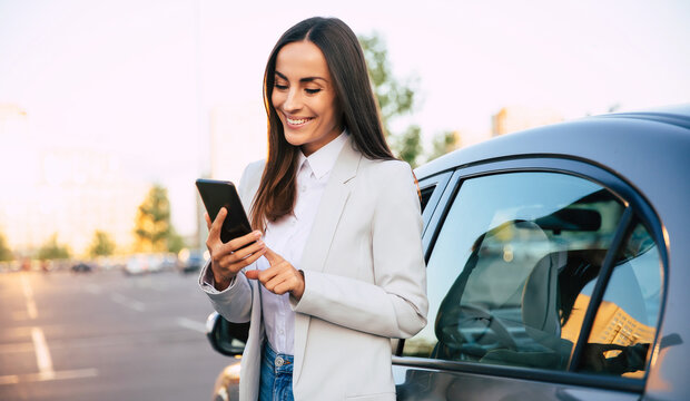 Successful smiling attractive woman in formal smart wear is using her smart phone while standing near modern car outdoors