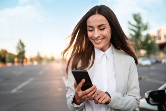 Successful Smiling Attractive Woman In Formal Smart Wear Is Using Her Smart Phone While Standing Near Modern Car Outdoors