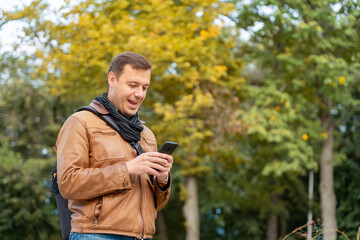Man with happy face checking phone standing in autumn park outdoors. Portrait of excited man holding smart phone in street.