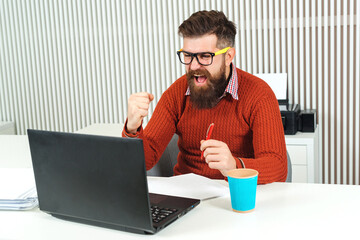 Happy bearded man working on laptop. Manager celebrating his success at workplace.