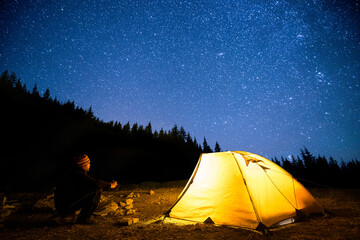 Traveler near the glowing camping tent on the background of the night forest and starry sky 