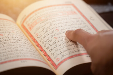 Fototapeta premium Quran (Koran) - close up of holy book of Muslims, with hand pointing ayat (Āyah), and shining light.