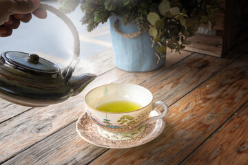 A woman pours green tea from an antique kettle into a warm porcelain cup laid on an old wooden floor.teacup with Chinese culture pattern