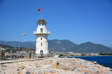 Fototapeta na wymiar white and blue lighthouse on bank near pier with pedestrian path and huge gray stones on sea and mountain background