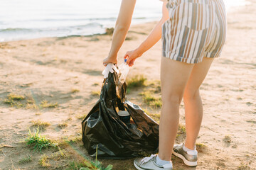 Young female volunteer satisfied with picking up trash, a plastic bottles and coffee cups, clean up beach with a sea. Woman collecting garbage. Environmental ecology pollution concept. Earth Day.
