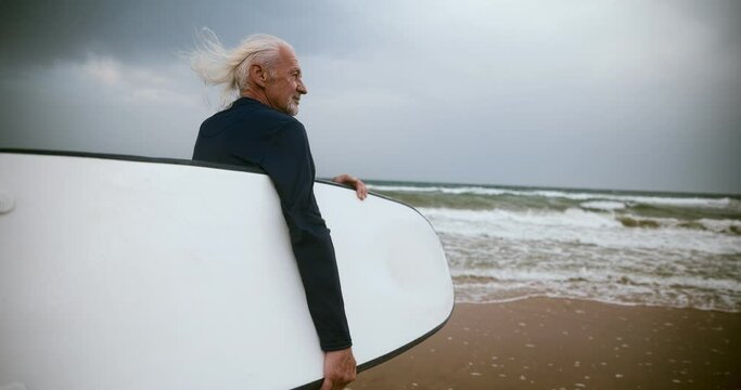 A gray-haired sporty old man goes surfing with a surfboard. An adult man surfs. 4 to Slow Motion
