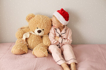 little beautiful girl in pink pajamas and santa hat is happy sitting on the bed with a teddy bear
