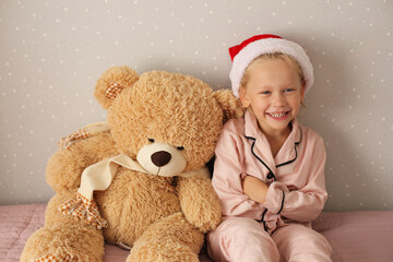 little beautiful girl in pink pajamas and santa hat is happy sitting on the bed with a teddy bear