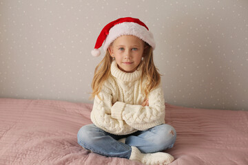 a beautiful little blonde girl is sitting at home on the bed in a winter sweater and a Santa claus hat new year Christmas