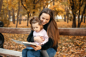 Young female woman babysitter and toddler baby girl read book in autumn park. Happy family mom and toddler outdoors in fall park - obrazy, fototapety, plakaty