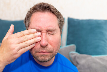 Obraz premium Middle aged 45s man holding hand over his aching eye at home on the couch.Closeup.