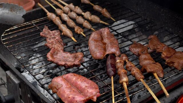 close-up of a grill with sausages, blood sausages and anticuchos of beef heart and chicken on a grill with charcoal fuming and seasoning with the skin of a corn with the hands in the day in 4k