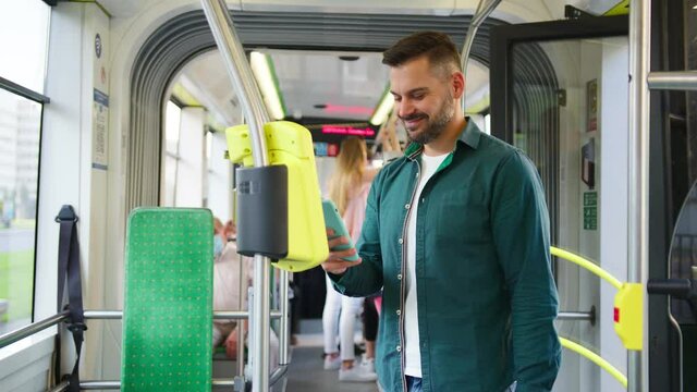 Young caucasian smiling man standing in the tram or bus as going somewhere, tapping and texting on his smartphone. Transportation and people concept