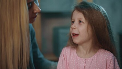 Smiling mother puts pill onto tongue of funny little daughter to cure disease on blurred background in semi-dark room at home close view slow motion