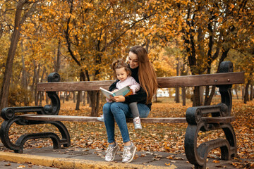 Young female woman babysitter and toddler baby girl read book in autumn park. Happy family mom and toddler outdoors in fall park - obrazy, fototapety, plakaty