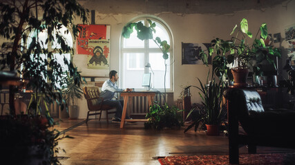 Fototapeta na wymiar Young Handsome Man Works on a Desktop Computer in Creative Agency in Authentic Loft Office. Renovated Stylish Decor with House Plants, Artistic Posters and Big Rounded Windows.