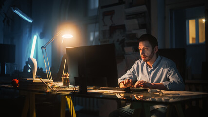Young Handsome Man Works on a Desktop Computer in Creative Agency in Dark Loft Office in the Evening. Renovated Stylish Decor with House Plants, Artistic Posters and Big Windows.