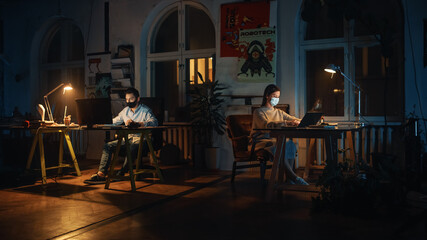 Two Stylish Employees Wearing Protective Face Masks while Working on Computers in Creative Agency in Loft Office. Colleagues Answer Emails and Manage Marketing Projects. Pandemic Covid-19 Concept.