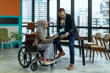Asian Muslim businesswoman sitting on wheelchair presenting business graph on paper to manager. Smiling group of diverse corporate colleagues in the modern office. Diversity or multicultural in office