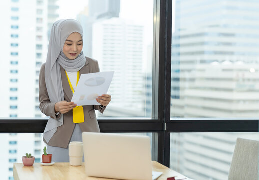 Asian Muslim Businesswoman In Hijab Head Scarf Planing Work With Paper Chart And Computer Laptop In The Modern Office