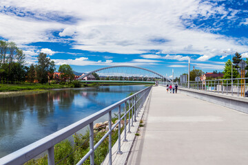 Walking path along the bank of the Koszarawa river. In the background, an arch-shaped bridge. A steel railing separating the sidewalk from the river..Żywiec, Poland