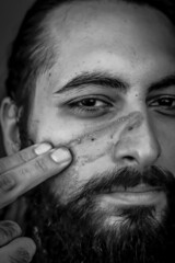 Tight black and white portrait of a bearded middle eastern man putting mud on his face	