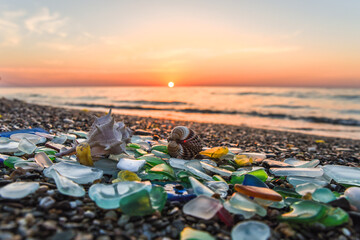 Natural polish textured sea glass and stones on seashore. Azure clear sea water with waves. Green, blue shiny glass with multi-colored sea pebbles close-up. Beach summer background, sunset and shells.