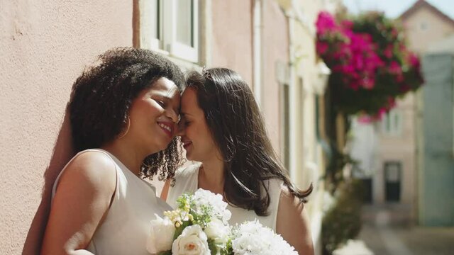 Smiling lesbian brides kissing and looking at camera. Medium shot of delightful African-American lovers posing for camera on sunny day. LGBT wedding, love, marriage concept