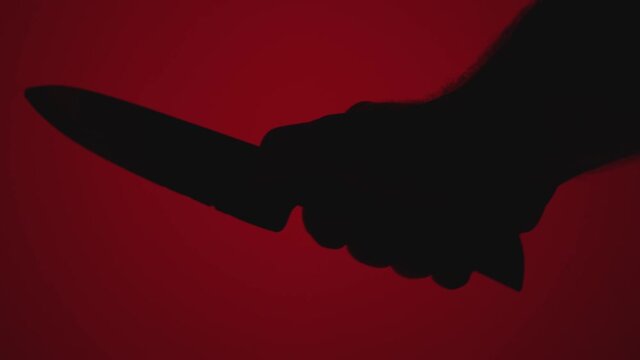 Close-up silhouette of a man's hand with a large knife against a red background. The concept of a homicidal maniac