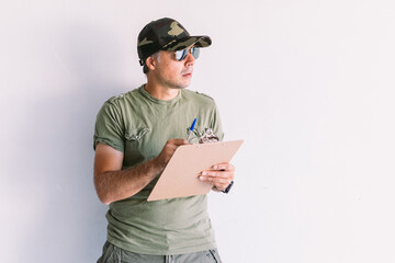 Military man wearing camouflage cap and sunglasses, writing in a report in a folder, on a white wall