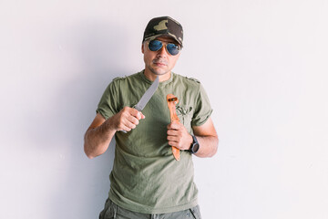Military man wearing camouflage cap and sunglasses, with a machete, on a white wall - obrazy, fototapety, plakaty