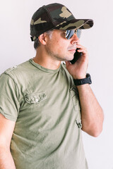 Military man with camouflage cap and sunglasses, talking on the phone, on a white wall