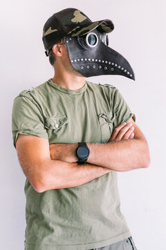 Military Man Wearing Camouflage Cap And Sunglasses, With Bubonic Plague Mask, On A White Wall