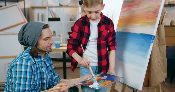 Attractive confident creative male artist holding colours palette during drawing picture together with cute interested boy in home workshop