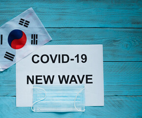 Fototapeta na wymiar On a blue wooden background is a sheet with the inscription: kovid-19 new wave, a medical mask and the flag of the Republic of Korea