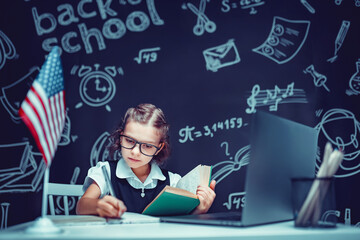 beautiful little schoolgirl sitting at desk and study online with laptop against black background with USA flag
