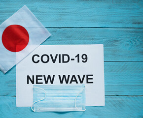 Fototapeta na wymiar On a blue wooden background are a sheet with the inscription: covid -19 new wave, a medical mask and a flag of Japan
