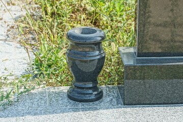 one empty black granite vase stands on a gray marble slab on a grave near a monument in a cemetery