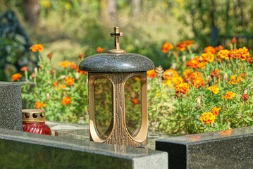 one big lamp with a cross stands on a black marble slab of a grave in a cemetery on the red flowers...