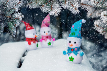 Three toy snowmen in the snow under a spruce tree during a snowfall. New Year and Christmas card