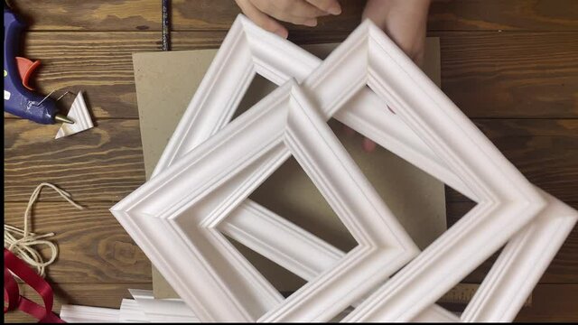 Women's hands show frames made of styrofoam of different sizes. Interior details made of moldings. Wooden table, hot glue and cardboard on background. Designer's workspace.