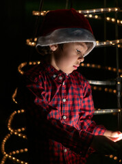 Fototapeta na wymiar Latino boy wearing a red santa claus hat looking concentrated down at his mobile phone with the light on his face and christmas tree lights on background. vertical