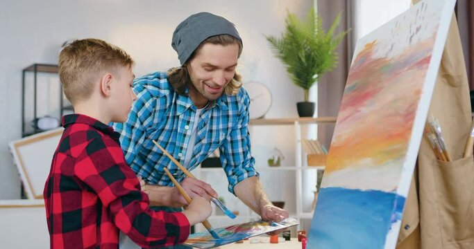 Good-looking smiling happy talented dad-artist having fun together his cute son during drawing picture using paintbrushes and paint palette in home studio