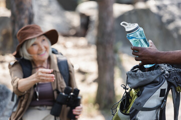African american traveler holding bottle of water near blurred wife in forest.