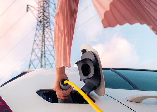 Female Hands Plugging In The Charger Into A Socket Of White Electric Car At A Charging Station Near An Electric Power Plant