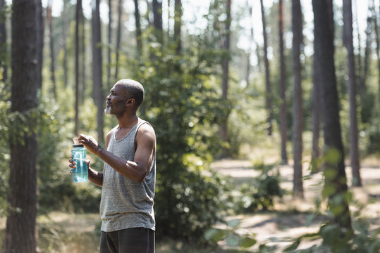 Side view of elderly african american sportsman holding sports bottle in forest.