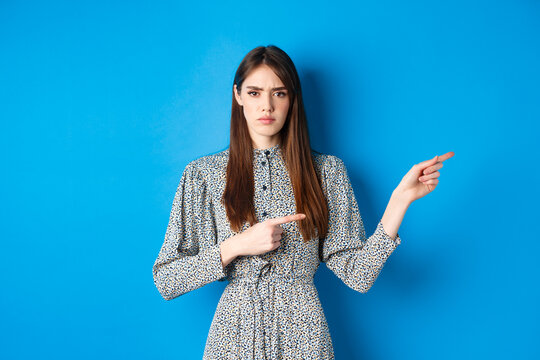 Angry Frowning Woman Pointing Fingers Right And Condemn Person, Feeling Disapointed, Standing In Dress On Blue Background