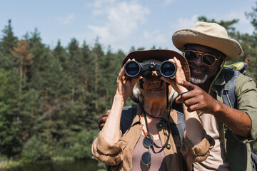 Smiling african american traveler pointing with finger near wife with binoculars in forest.