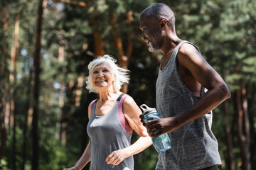 African american sportsman holding sports bottle while jogging with wife in forest.